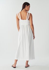 Hvid maxikjole med et tætsiddende livstykke og justerbare stropper, der har en rynket detalje på ryggen og en flydende nederdel. Bruges med flade sandaler.