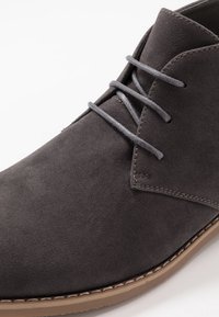 Chaussure en daim gris avec une finition texturée, présentant des lacets gris et une semelle en caoutchouc beige contrastante, soulignée par un accent cousu le long du bord.