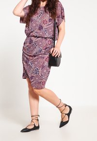Vestido púrpura con estampado, de mangas cortas, diseño drapeado y un bolso bandolera negro. Unos zapatos negros planos con tiras en el tobillo completan el conjunto.