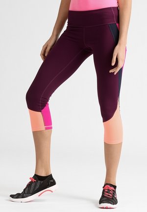 Mujer vistiendo leggings cortos con bloques de color púrpura, rosa y melocotón, con zapatillas deportivas negras y rosas, de pie con las piernas ligeramente separadas.