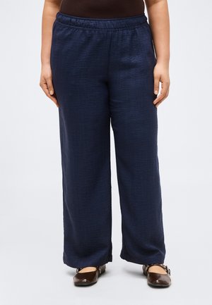 Personne portant un pantalon ample bleu marine avec une ceinture élastique, un haut marron foncé et des chaussures plates marron avec des brides à la cheville.