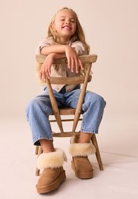 Stivali di camoscio marrone con polsini di pelliccia morbida color crema, abbinati a jeans di denim chiaro e un cardigan beige in maglia, seduti su una sedia di legno.