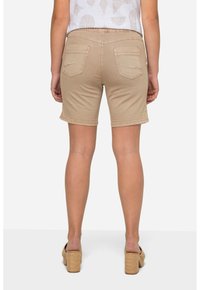 Shorts in denim beige con tasche posteriori, orli rifiniti e un design aderente. Il modello indossa un top bianco a fantasia e sandali con tacco marrone chiaro.