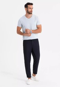 Vaaleansininen V-kaula-aukkoinen t-paita, tummansiniset joustavapohjaiset housut, valkoiset liukuvat kengät; malli seisoo kädet ristissä, minimaalinen tausta.