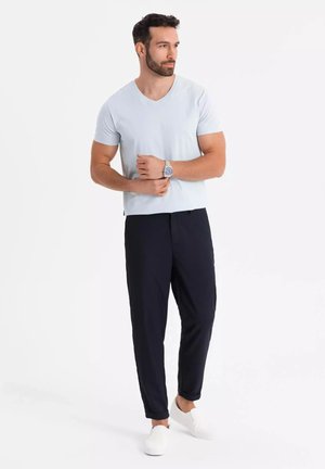 Lyseblå V-hals t-shirt, mørk navy elastiske bukser, hvide slip-on sko; model står med armene krydset, minimal baggrund.