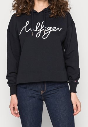 Kobieta w czarnej bluzie z kapturem z logo napisowym "Hilfiger" oraz ciemnoniebieskich dżinsach, ręce luźno opuszczone wzdłuż ciała.