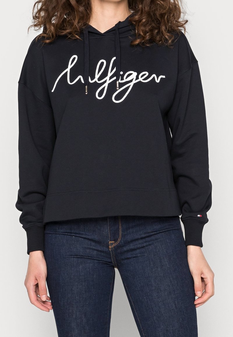 Femme portant un sweat à capuche noir avec le logo script "Hilfiger" et un jean bleu foncé, les mains détendues le long du corps.