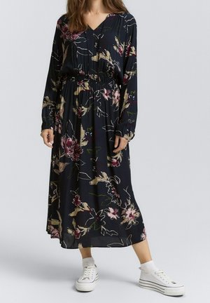 Femme portant une robe midi noire à manches longues avec un motif floral, associée à des chaussettes blanches et des baskets compensées blanches, sur un fond uni.