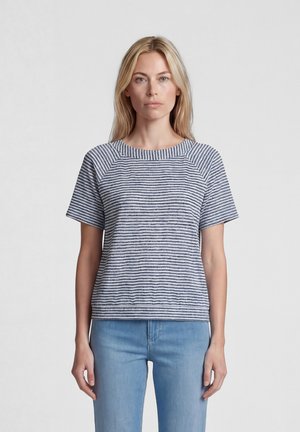 Blonde woman wearing a short-sleeve navy and white striped top and light blue jeans, standing against a plain gray background.