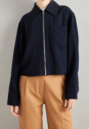 Personne portant une veste zippée bleu marine avec col et poche poitrine, associée à un pantalon taille haute couleur camel sur un fond neutre.