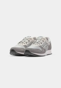 Light gray sneakers with a mix of mesh and suede materials, featuring a rounded toe, laces, and a patterned sole. Logo on the tongue.
