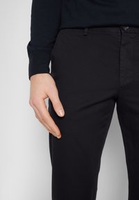 Svarta bomullsbukser med en slät struktur, slim fit, framfickor och knappstängning. En hand vilar avslappnat på lårområdet.