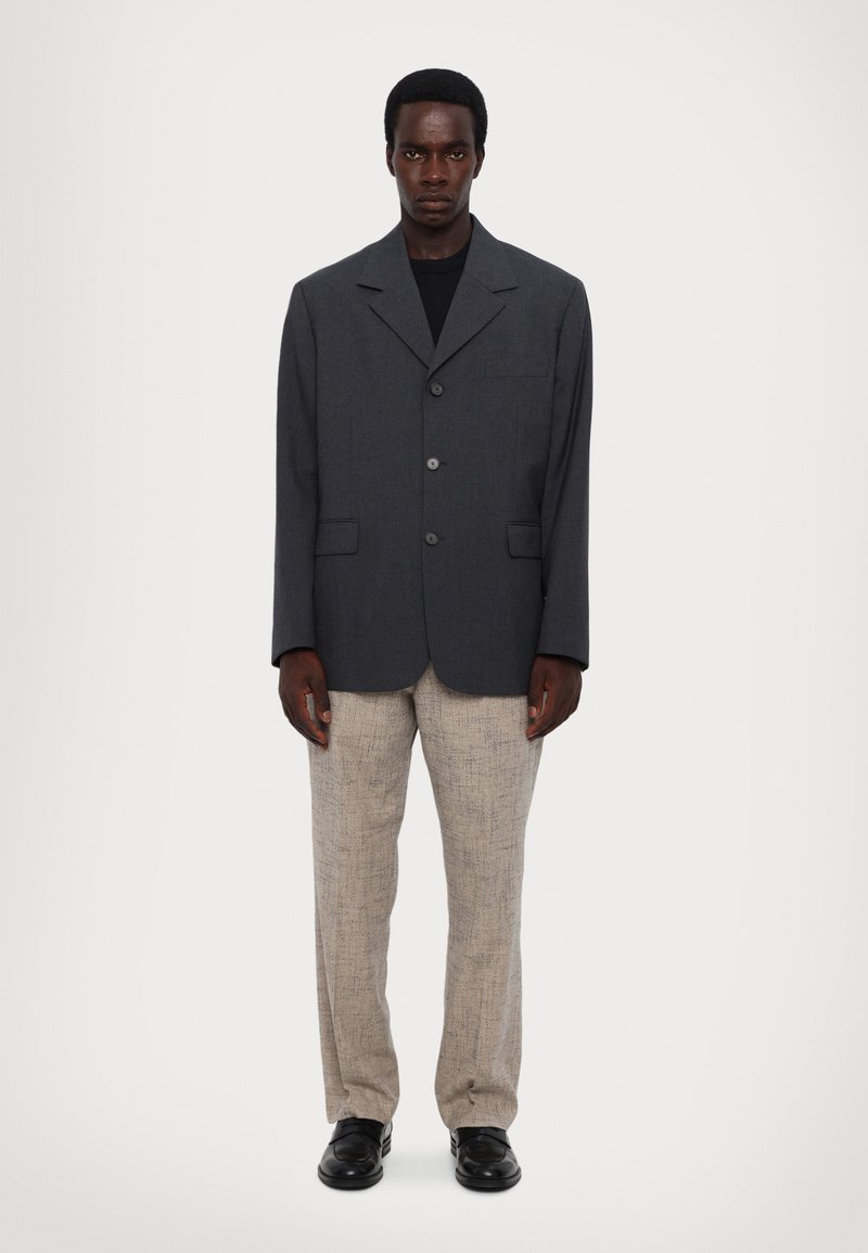 Tumehall oversized bleiser kahte nööbi ja rinnakoti kanssa, millele on lisatud beežid tekstuuriga püksid ja mustad kingad.