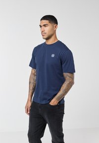 Man with tattoos wearing a navy blue t-shirt and black jeans, standing with one hand in pocket against a plain background.