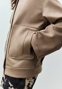 Veste beige, douce, avec texture, dotée d'une fermeture à boutons, d'une grande poche plaquée et d'une coupe décontractée, associée à un pantalon sombre à motifs.