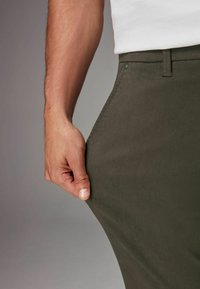 Pantalones verde oliva hechos de una tela lisa y texturizada, con un primer plano de una mano sosteniendo la cinturilla, mostrando el diseño y el ajuste.