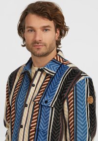 Hombre con barba clara y cabello castaño ondulado, vistiendo una chaqueta a rayas multicolores con un bolsillo en el pecho abotonado y una camisa beige debajo.