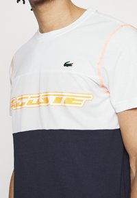 Kortärmad t-shirt med vita mesh- och marinblå bomullspartier, med orange sömmar och en gul "Lacoste"-logotyp på bröstet.