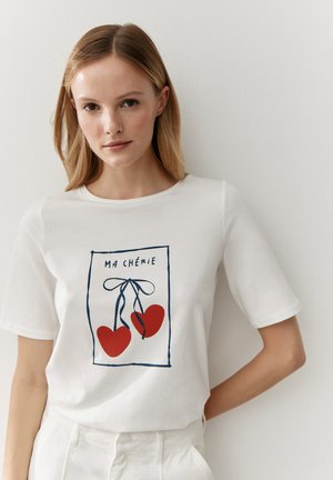 Junge Frau trägt ein weißes T-Shirt mit einem rot gezeichneten Herzen in Form von Kirschen und dem Schriftzug "Ma Chérie", steht vor einer einfachen hellen Wand.