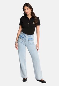 Sort polo-shirt med knapper og et badge, parret med lyseblå vide bukser med to forlommer. Sorte flade sko fuldender outfittet.
