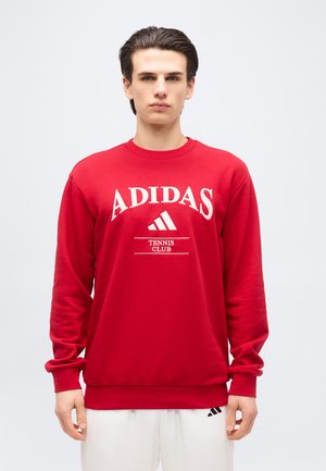 Νεαρός άνδρας που φοράει κόκκινο φούτερ Adidas με το κείμενο και το λογότυπο «Tennis Club», συνδυασμένο με λευκό παντελόνι, σε απλό φόντο.