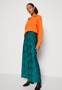 Suéter de punto naranja con cuello, combinado con una falda teal con pliegues y rayas verticales. El modelo lleva botas negras hasta el tobillo.