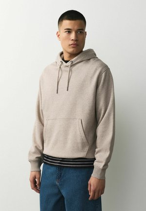 Young man wearing beige hoodie with front pocket and blue jeans, standing against plain light gray background, looking right.