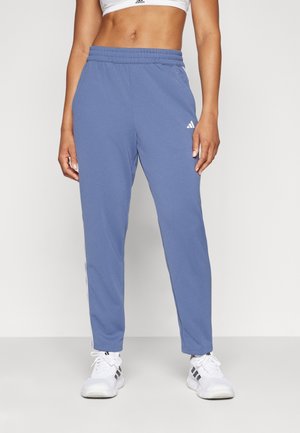 Kobieta ubrana w niebieskie spodnie dresowe Adidas z elastycznym pasem, biały sportowy biustonosz i białe trampki Adidas z czarnymi paskami.