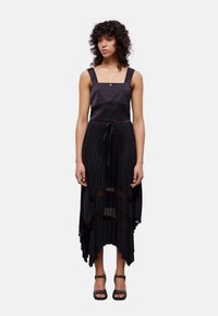 Fekete ujjatlan ruha négyzet alakú nyakkivágással, elöl gombokkal, redős szoknyával és magas-alacsony szegélyű kialakítással. Fekete szandállal viselve.