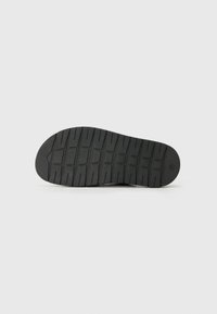 Semelle de chaussure noire avec des sculptures profondes et des motifs rectangulaires, vue par le dessous sur un fond gris clair uni.