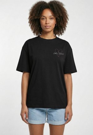Voksen kvinde med krøllet hår iført en sort oversize T-shirt med pink hjerteprint og lyseblå oprullede denimshorts, stående oprejst.