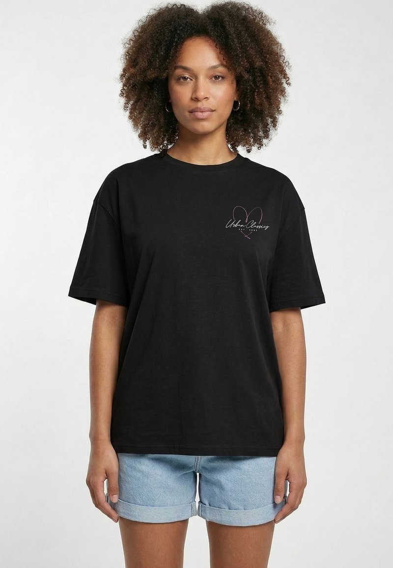Femme adulte aux cheveux bouclés portant un T-shirt noir oversize avec un motif cœur rose et un short en jean bleu clair retroussé, debout.