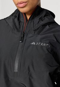 Die schwarze Terrex-Jacke verfügt über einen hohen Kragen, einen frontalen Reißverschluss mit roten Akzenten und einem gestickten Logo. Das Material scheint leicht und wasserdicht zu sein.
