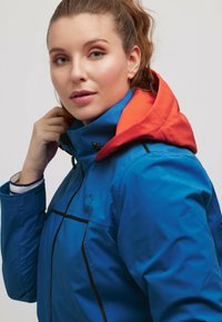 Blauwe waterdichte jas met oranje capuchon, zwarte accenten en een hoge kraag; beschikt over een merklogo en stevige sluitingen.