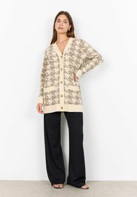 Beige and brown houndstooth cardigan with buttons, v-neck, and pockets. Paired with loose black trousers, featuring a smooth texture and wide legs.