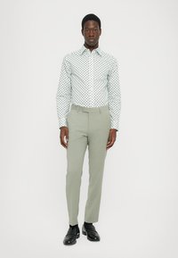 White shirt with black polka dots, long sleeves, buttoned collar; light green tailored trousers; black shoes; neutral background.