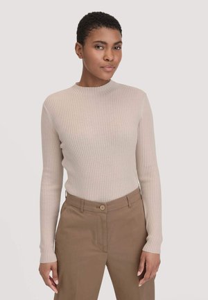 Jonge vrouw met kort haar, gekleed in een beige geribbelde lange mouwtop en lichtbruine hoge taillebroek, staande tegen een effen achtergrond.