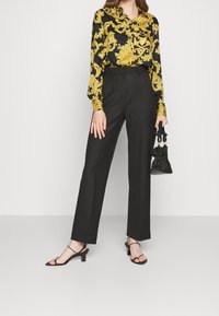 Kobieta w czarnych spodniach i czarnych sandałach na wysokim obcasie z paskami, trzymająca małą błyszczącą czarną torebkę, w zestawieniu z czarną bluzką w żółte kwiaty.