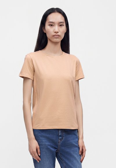Femme aux longs cheveux noirs portant un t-shirt beige uni à manches courtes et un jean bleu, debout sur un fond blanc uni.