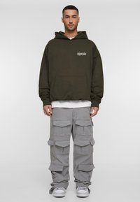 Olive green hooded sweatshirt with a front pocket, layered over a white tee. Paired with gray cargo pants featuring multiple pockets. White sneakers.