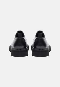 Zapatos de cuero negro con un acabado suave, punta redondeada y suela de goma gruesa. Cuentan con lengüetas en la parte trasera para facilitar su colocación.