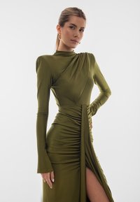 Olivově zelené šaty s dlouhými rukávy, strukturovaným detailem na rameni, bočními nabíráními a vysokým rozparkem ke stehnům. Hladká, elastická tkanina. Viditelné zlaté náušnice.