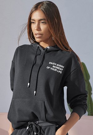 Zwarte hoodie met trekkoord, voorzak en witte tekst "GENIET VAN ELK MOMENT VAN JE LEVEN" op de borst, gemaakt van zachte stof.