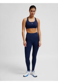 Granatowy stanik sportowy i legginsy z wysokim stanem wykonane z elastycznego materiału. Posiada minimalistyczne logo na staniku, zestawione z białymi i niebieskimi butami sportowymi.