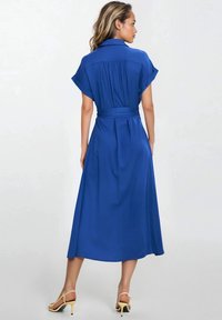 Niebieska sukienka do kolan z krótkim rękawem i paskiem w talii. Gładka tekstura materiału, plisowania z tyłu, noszona z żółtymi sandałami na obcasie.