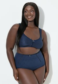 Uśmiechnięta kobieta z długimi prostymi włosami, ubrana w dopasowany granatowy biustonosz i wysokie majtki na białym tle.