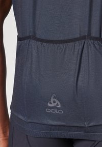 T-shirt de sport bleu foncé en tissu léger, comportant une section froncée à la taille et un logo discret en bas.