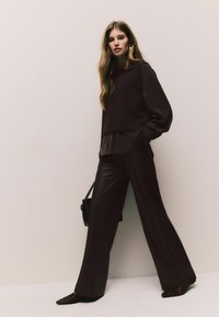 Brown pullover sweater over a dark shirt, paired with wide-leg brown leather pants and animal-print shoes. Black handbag as an accessory.