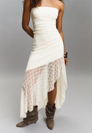 Trägerloses weißes asymmetrisches Kleid mit Spitzenüberzug, kombiniert mit lässigen braunen Wildlederstiefeln und einem auffälligen Armband am rechten Handgelenk.