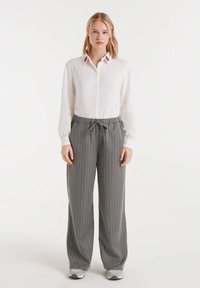 Joven mujer vestida con una camisa blanca de botones con bordado floral en el cuello, pantalones de pinzas grises de pierna ancha y zapatillas blancas, de pie.